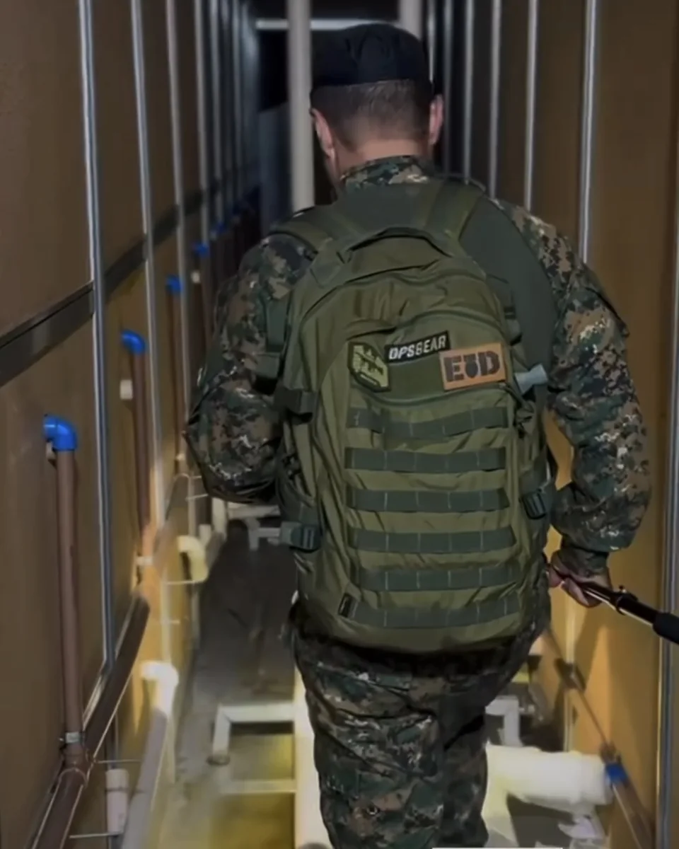 Operador de costas com mochila tática verde OPSGEAR e patches, caminhando em corredor